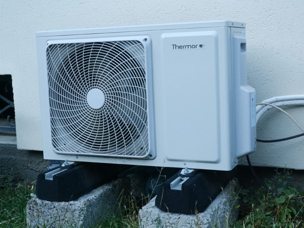 photo d'une unité extérieure de climatiseur de phase. La climatisation est de la marque thermor (marque française). L'unité extérieure et surélever par des blocs en béton puis sur les calle noire, ça ressemble à de la mousse. Dessous il y a de l'herbe et derrière un mur crépi de couleur crème.