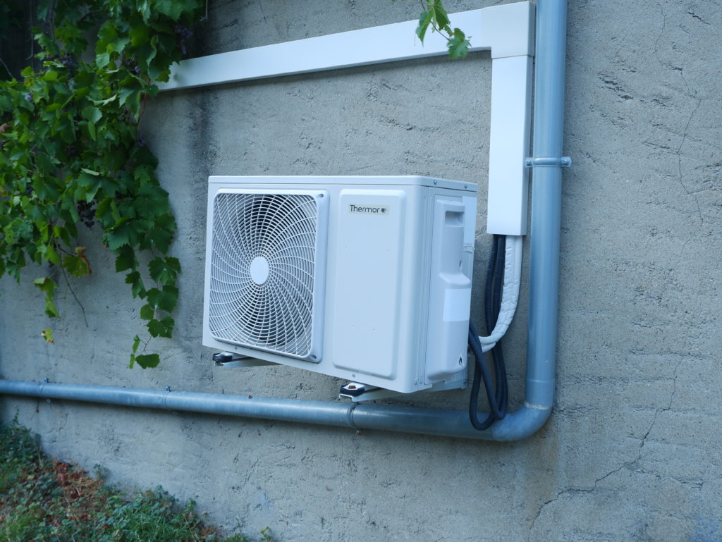 une unité climatisation extérieure fixer à un mur de couleur gris. on peut y voir une descente de chenaux qui passe le côté droit de l'uniter climatisation puis dessous l'unité. Sur la gauche de l'unité climatisation, toujours sur le mur il y a un pied de vigne.