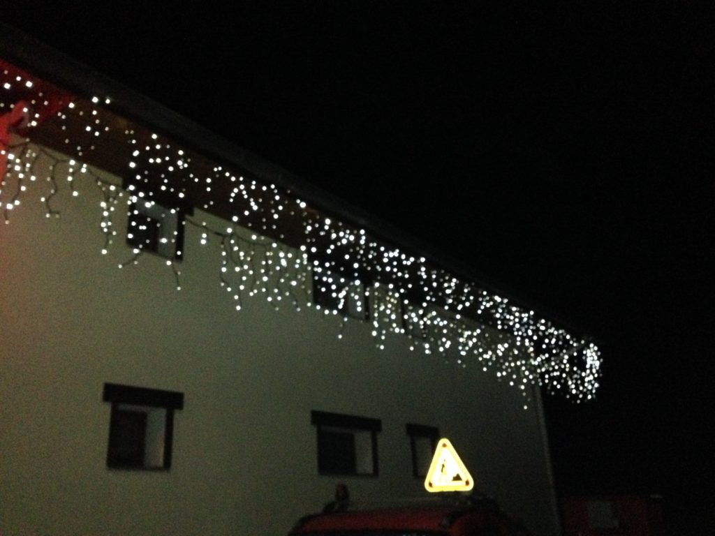 il fait nuit. on voit le côté d'une maison, il y a quelques petites fenêtres. Sur le bord du toit, au niveau des goutières, il y a une guirlande lumineuse de couleur blanche froide qui est éclairée, c'est une guirlande de Noël. il y a différentes tailles de fils lumineux qui pendouillent, c'est une guirlande à tablier chute d'eau.