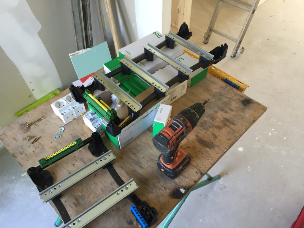 Dans un chantier, il y a une sorte de table/établit de chantier en bois avec des pieds en métal de couleurs vertes. Sur cette table il y a une viseuse, des bloque de prise de courant ainsi de ces cartons et tout le nécessaire pour la création d'un tableau électrique.