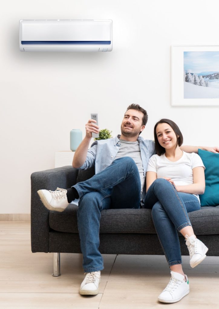 en premier plan de cette photo, nous voyons un jeune couple tout sourire, assis sur un canapé gris. monsieur a un bras autour des épaules de madame. son autre bras est plié et tiens dans sa main une télécommande. en arrière-plan il y a un mur blanc avec un tableau sur la droite et une unité de climatisation blanche sur la gauche. tout nous laisse penser que le jeune couple est heureux, confortablement installer et que monsieur regle la température de la climatisation pour avoir encore plus de confort