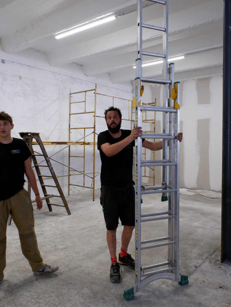 SALOMON Hervé qui bouge une echelle. on voit un morceau de l'apprentis sur la gauche de la photo et un echafaudage a l'arriere plan. ils installent des luminaire dans un entrepot.